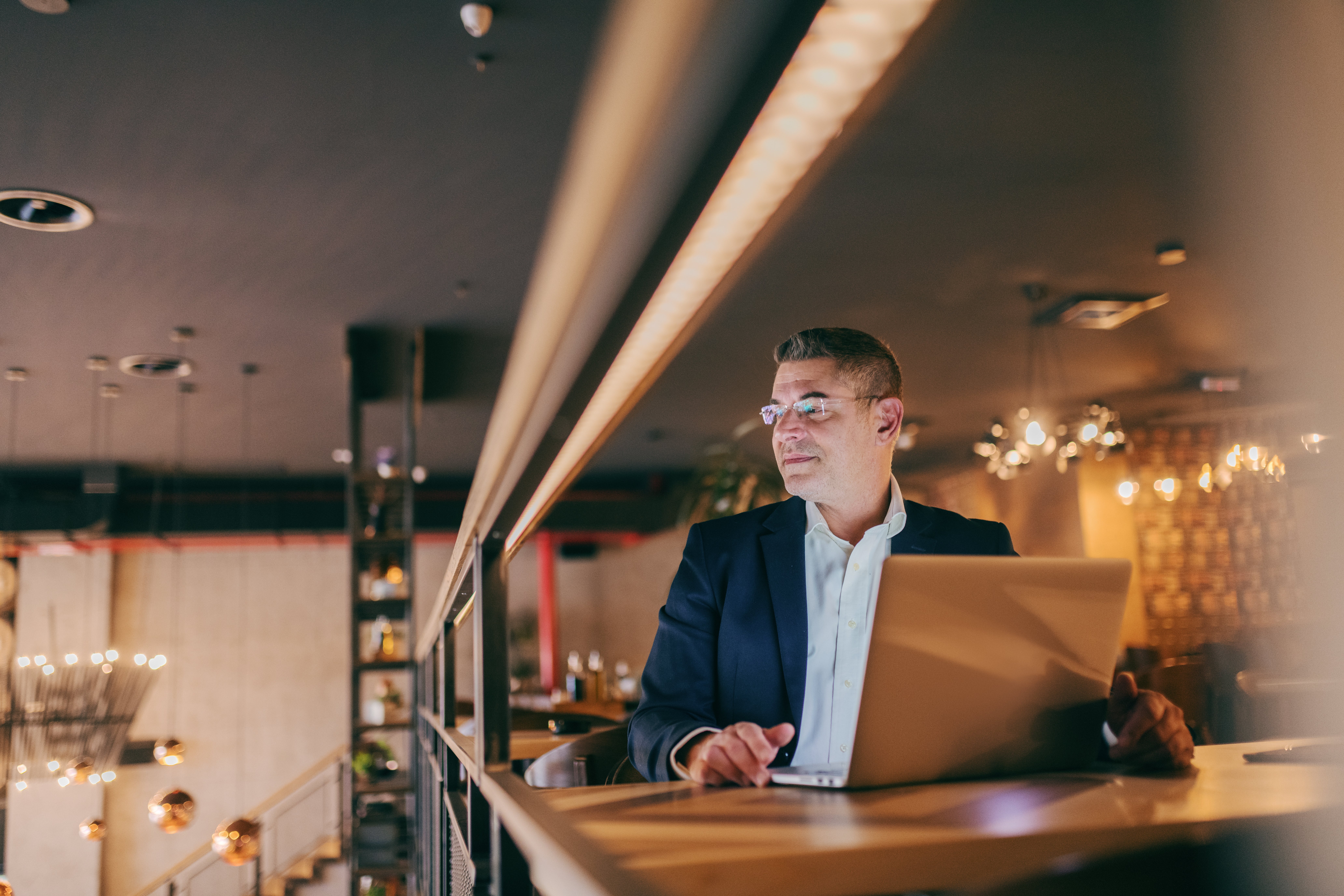 A man sits at a table in a restaurant and looks away from his laptop screen