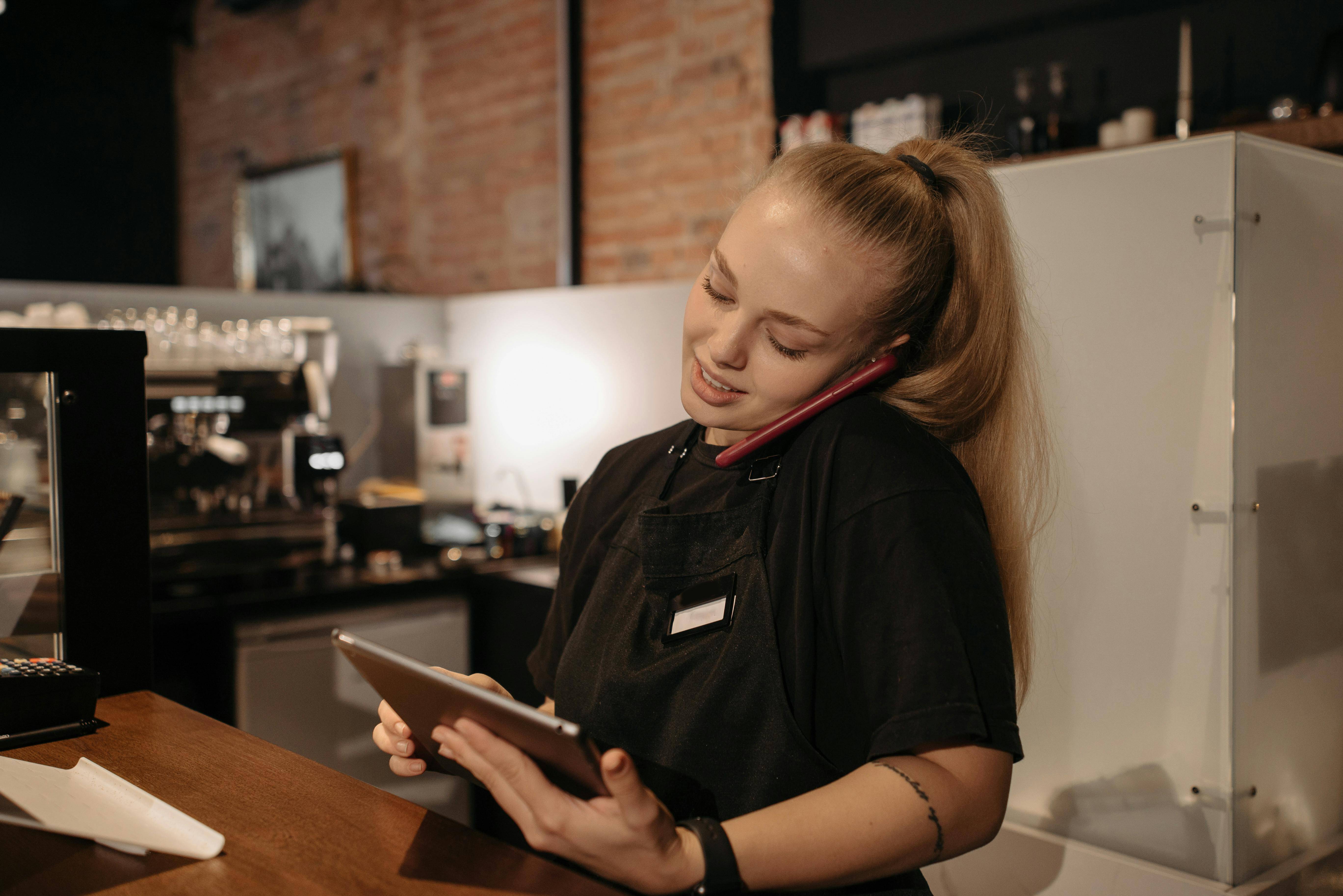 Mitarbeiterin telefoniert und arbeitet gleichzeitig auf einem Tablet hinter der Theke eines Restaurants